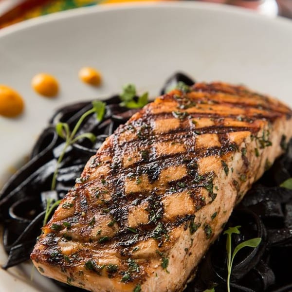 Grilled salmon with black fettuccine pasta, garnished with herbs and orange sauce dots.