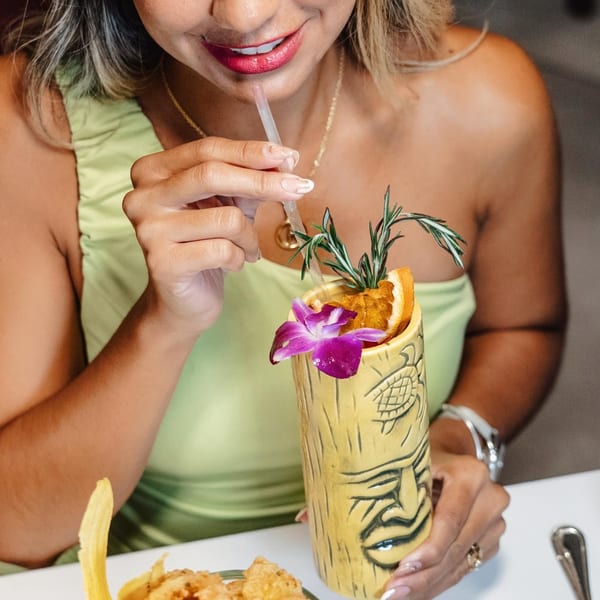 A woman sips a vibrant cocktail from a tiki mug, garnished with rosemary, orange, and an orchid.