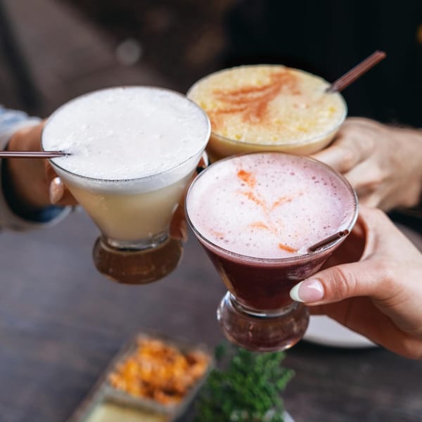 Three hands holding up different colored Pisco Sours, a classic Peruvian cocktail, for a toast.