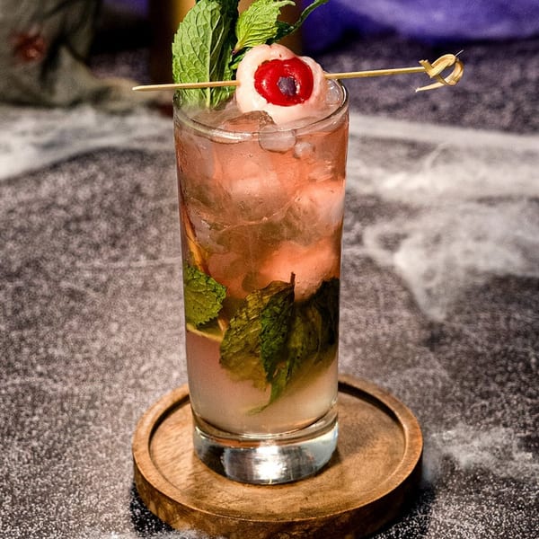 A Peruvian cocktail with mint, ice, and a lychee-cherry garnish, on a wooden coaster.