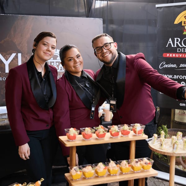 Three smiling servers in maroon jackets stand behind a display of small appetizers.