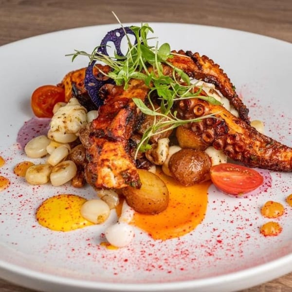 Peruvian grilled octopus with baby potatoes, corn, cherry tomatoes, and microgreens on a white plate.