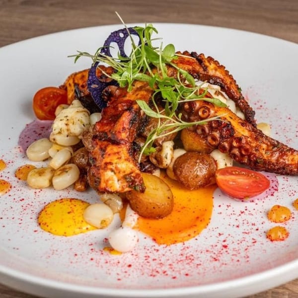 Grilled octopus with baby potatoes, cherry tomatoes, pearl onions, and colorful sauces on a white plate.