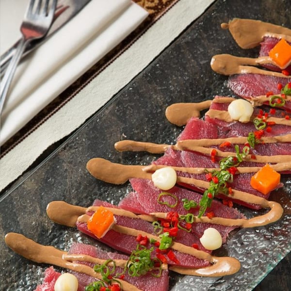Peruvian tuna tiradito with aji amarillo sauce, rocoto, chives, and white corn on a dark plate.