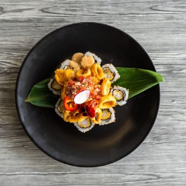 Peruvian sushi rolls topped with fried calamari, salsa criolla, and a radish slice on a black plate.