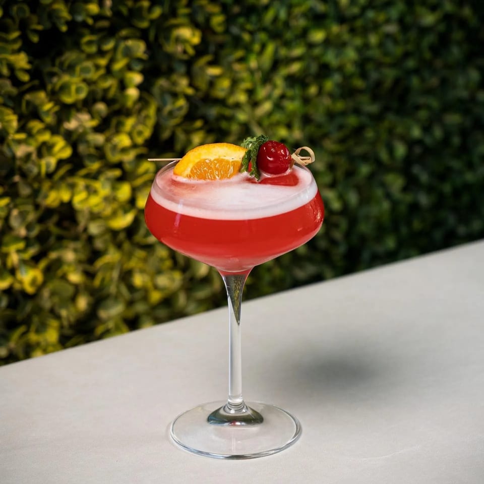 A vibrant red cocktail with white foam, garnished with an orange slice, cherry, and mint, against a green foliage background.