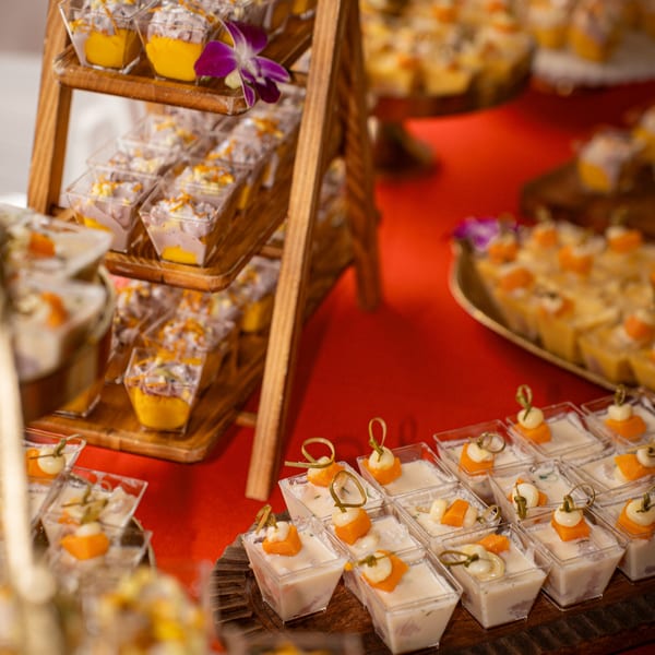 Dessert cups with yellow and white layers, topped with fruit, on a tiered wooden stand and red tablecloth.
