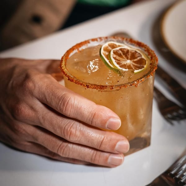 A hand holding a Peruvian cocktail with a chili-salted rim and dried lime garnish.