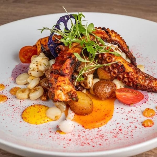 Grilled octopus with potatoes, corn, cherry tomatoes, and sauces on a white plate.