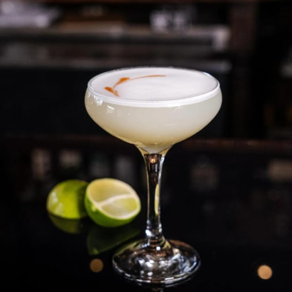 A Pisco Sour cocktail with foam and bitters, garnished with lime halves on a dark bar.