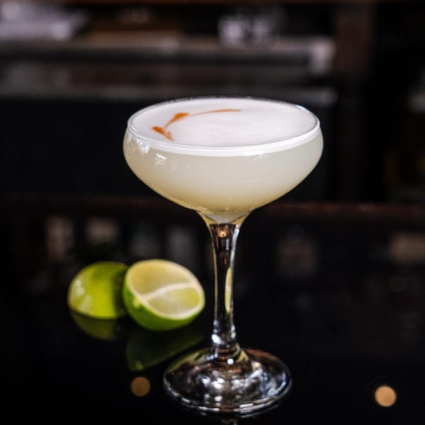 A Peruvian Pisco Sour cocktail with foam art and two lime halves on a dark surface.