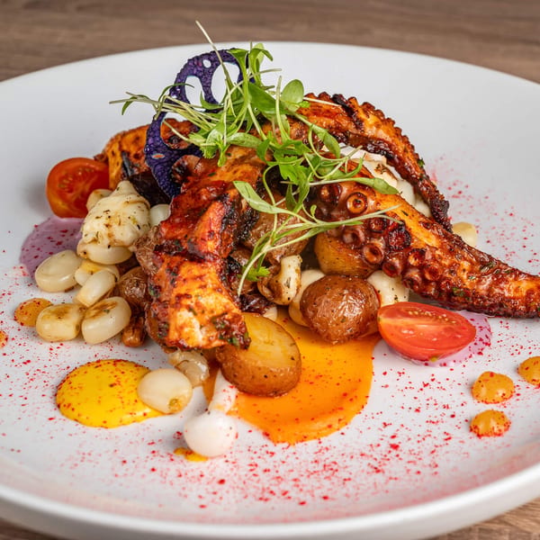 Peruvian grilled octopus with potatoes, corn, cherry tomatoes, and microgreens on a white plate.