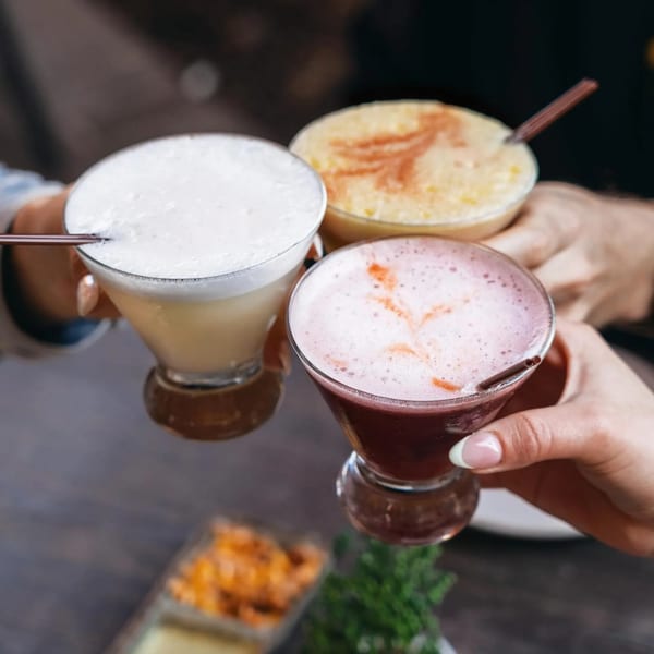 Three hands holding different colored Pisco Sours, including classic, chicha morada, and passion fruit.