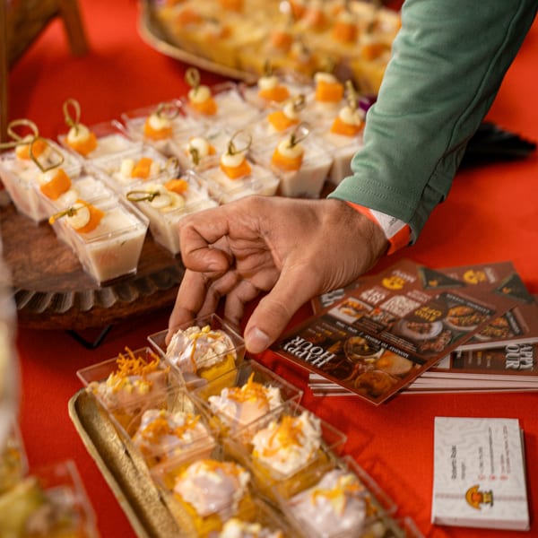 A hand reaches for a small dessert cup with white cream and orange topping on a red table.