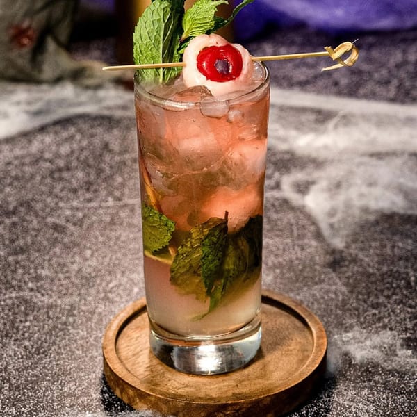 A Peruvian cocktail with mint and a lychee-cherry garnish, served on a wooden coaster.